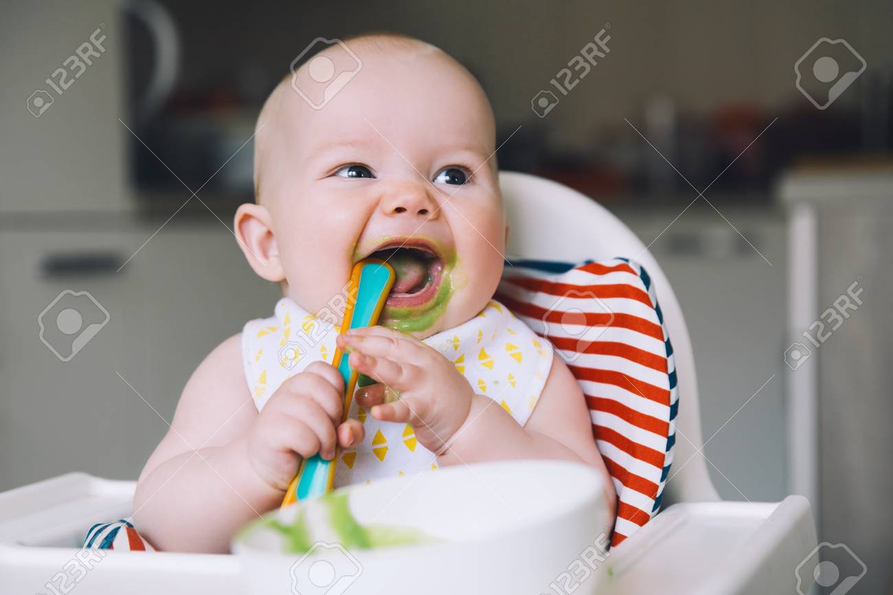 Feeding. Messy smiling baby eating with a spoon in high chair. Baby's first solid food. Mother feeding little child with spoon of puree. Daily routine. Finger food. Healthy child nutrition. - 100136351 Feeding. Messy smiling baby eating with a spoon in high chair. Baby's first solid food. Mother feeding little child with spoon of puree. Daily routine. Finger food. Healthy child nutrition. - 100136351