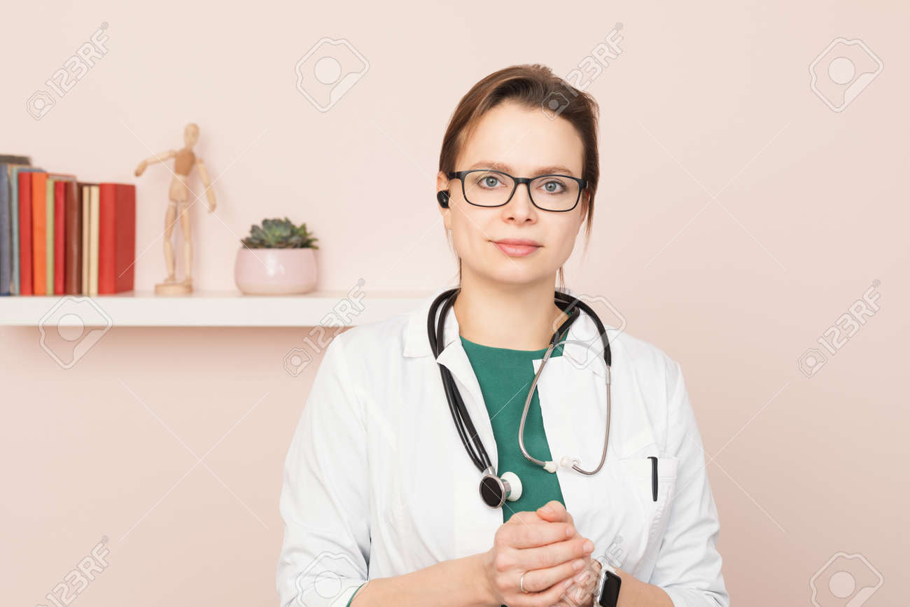 Portrait of confident young female doctor wearing white coat with stethoscope and earpiece standing in front of a camera at her home office performing online consultation - 164061906 Portrait of confident young female doctor wearing white coat with stethoscope and earpiece standing in front of a camera at her home office performing online consultation - 164061906