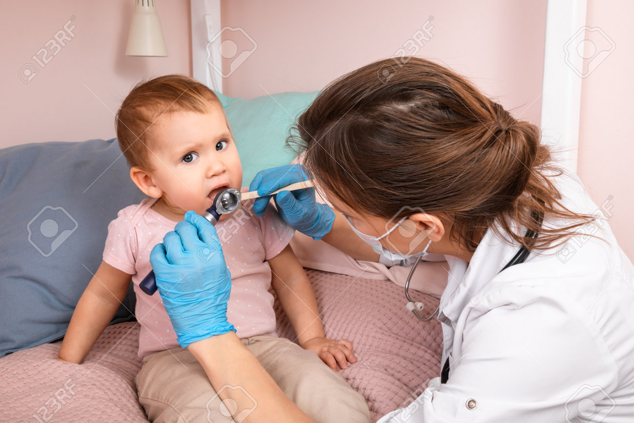 Pediatrician examines mouth of a baby girl at home during quarantine. Doctor using wooden tongue depressor and torch - 165037434 Pediatrician examines mouth of a baby girl at home during quarantine. Doctor using wooden tongue depressor and torch - 165037434