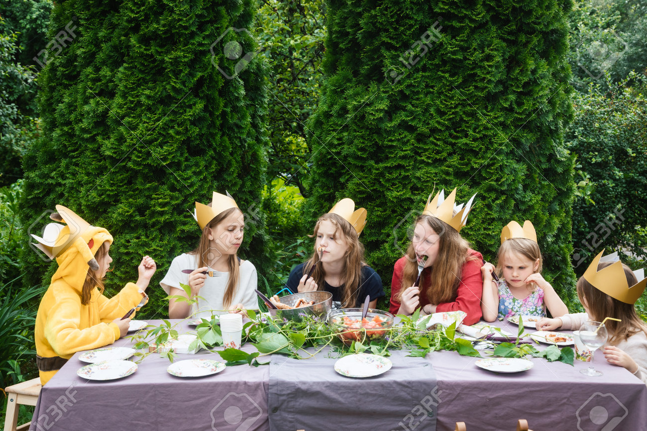 Children wearng paper crowns sitting by decorated table eating grilled sausages celebrating birthday party in a green garden - 167165492 Children wearng paper crowns sitting by decorated table eating grilled sausages celebrating birthday party in a green garden - 167165492