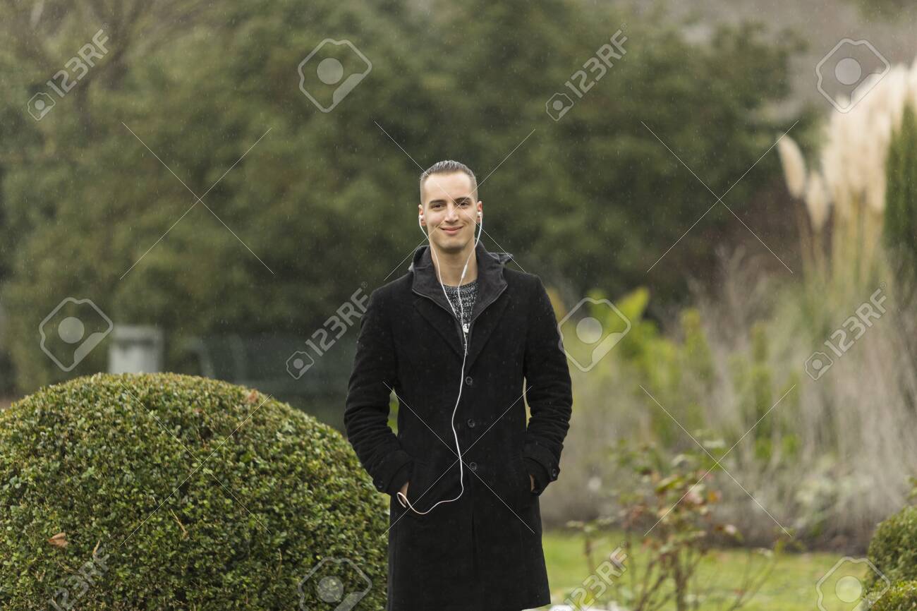 Young Man Wearing Coat and Listening to Earphones Posing in Garden - 143965966 Young Man Wearing Coat and Listening to Earphones Posing in Garden - 143965966