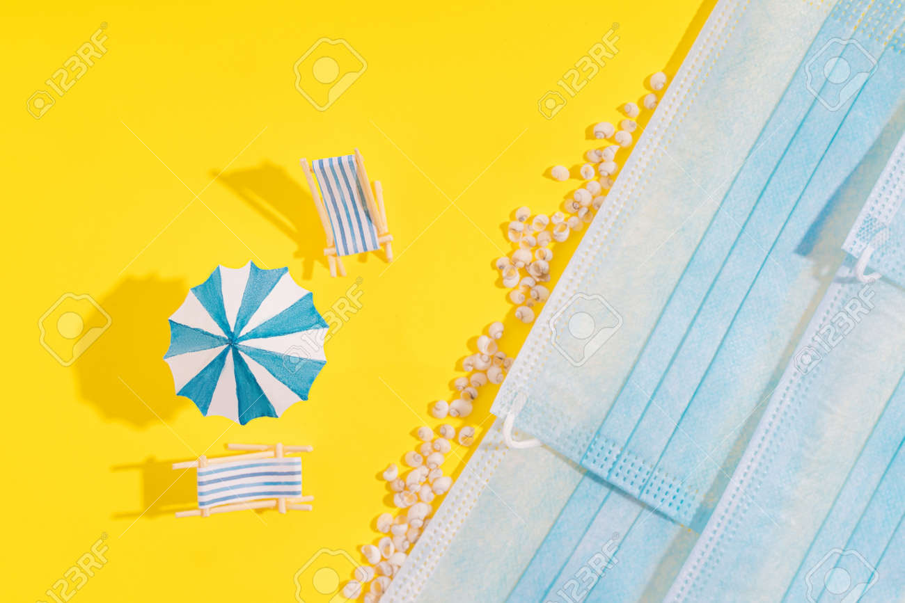 Summer vacation concept during pandemic. Medical masks are depicted as sea and yellow background as sand. Two sun loungers, an umbrella and small seashells standing on yellow background. - 169603055 Summer vacation concept during pandemic. Medical masks are depicted as sea and yellow background as sand. Two sun loungers, an umbrella and small seashells standing on yellow background. - 169603055