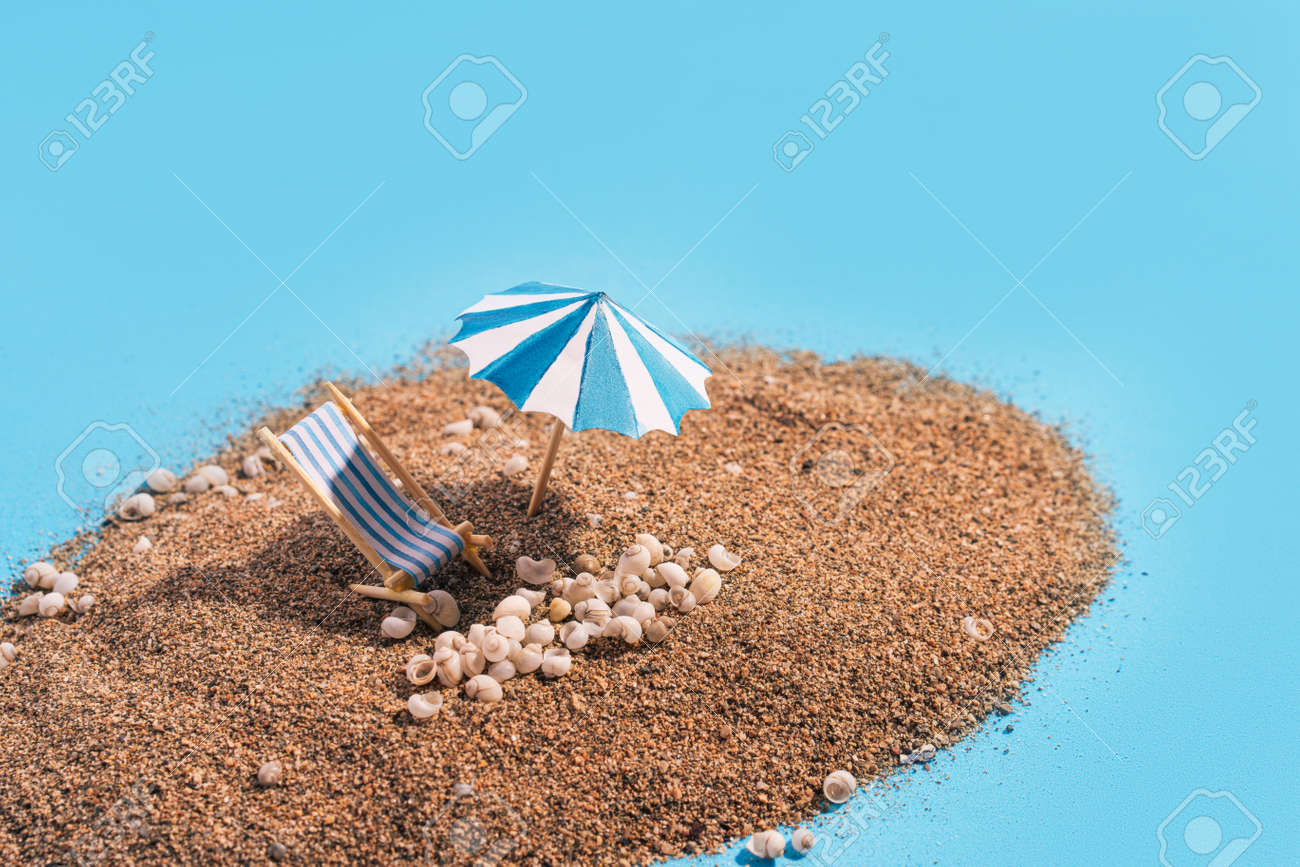 Summer vacation concept during pandemic. Slide of sand on blue background like the sea. Deck chair, umbrella and small seashells on the sand. - 169603056 Summer vacation concept during pandemic. Slide of sand on blue background like the sea. Deck chair, umbrella and small seashells on the sand. - 169603056