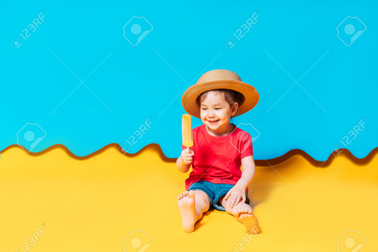 Asian girl in a straw hat licks ice cream. The blue background shows the sky, and the yellow background shows the sand. - 169603104 Asian girl in a straw hat licks ice cream. The blue background shows the sky, and the yellow background shows the sand. - 169603104