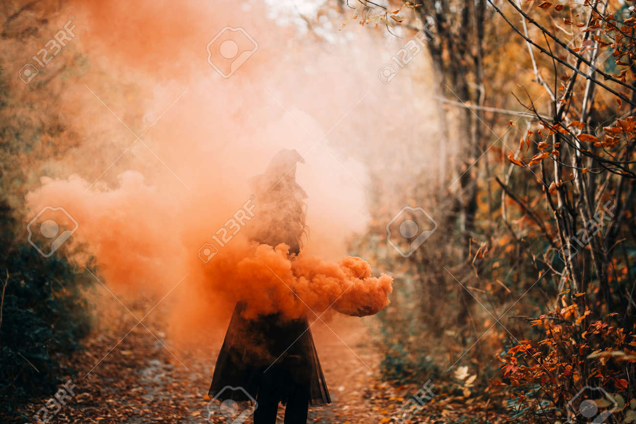 Girl dressed as witch in orange smoke on forest background. - 169694346 Girl dressed as witch in orange smoke on forest background. - 169694346