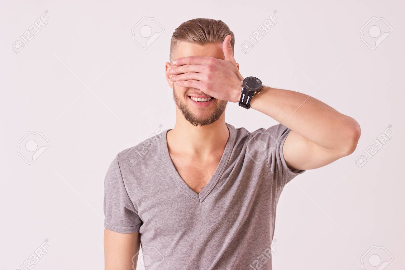 145235378-studio-shot-of-cheerful-young-man-covering-his-eyes-with-hand-and-smiling-while-isolated-on-white-ba.jpg