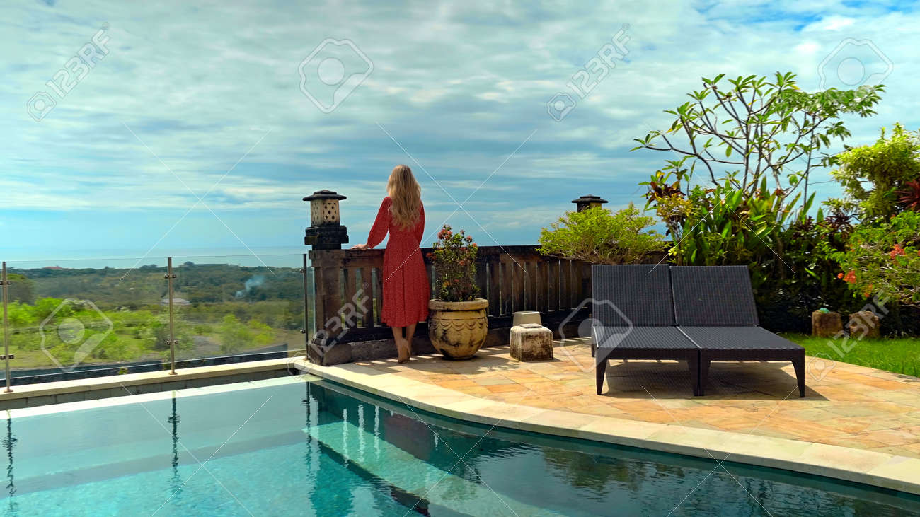 Young long-haired girl walking in the backyard with a green garden, pool with blue clean water and blue sky in a beautiful long red dress - 171458560 Young long-haired girl walking in the backyard with a green garden, pool with blue clean water and blue sky in a beautiful long red dress - 171458560