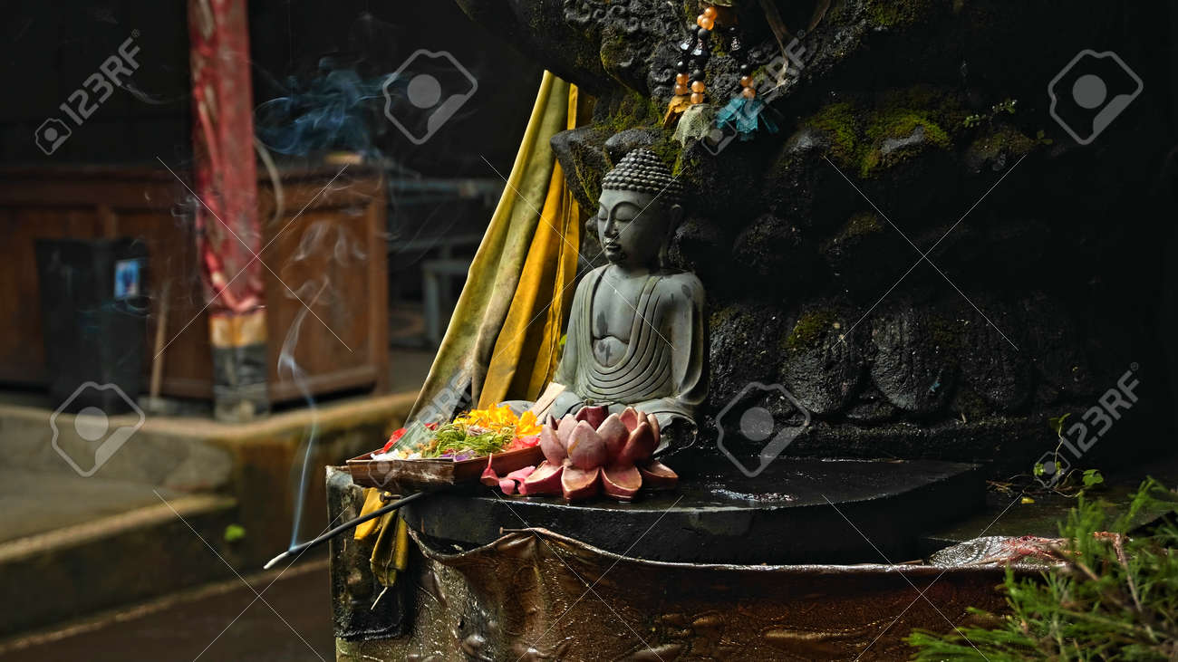 An old stone Budist statue sitting in a lotus pose covered with moss with a wreath of yellow flowers on its neck standing on the street with green plants on the background - 171458567 An old stone Budist statue sitting in a lotus pose covered with moss with a wreath of yellow flowers on its neck standing on the street with green plants on the background - 171458567