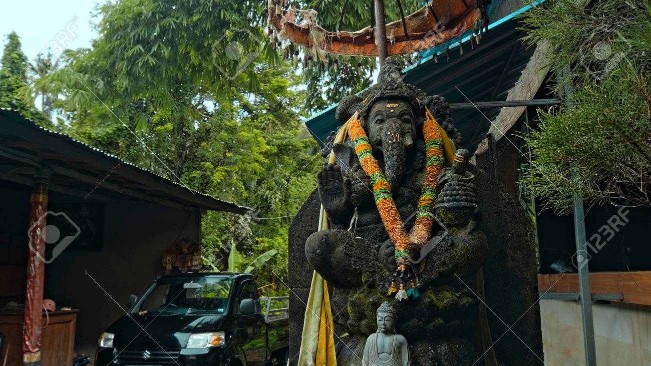 An old stone statue of Ganesha covered with moss with a wreath of yellow flowers on his neck standing on the street with green plants on the background - 171544658 An old stone statue of Ganesha covered with moss with a wreath of yellow flowers on his neck standing on the street with green plants on the background - 171544658