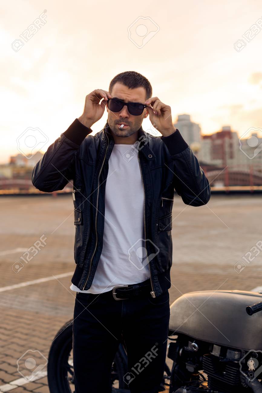 ひげと黒のバイカー ジャケット正しいファッション サングラス日没でクラシックなスタイルのカフェ レーサー バイク近くタバコを喫煙で口ひげを持つハンサムなライダー男のクローズ アップ 残忍な都会のライフ スタイルを楽しい の写真素材 画像素材 Image
