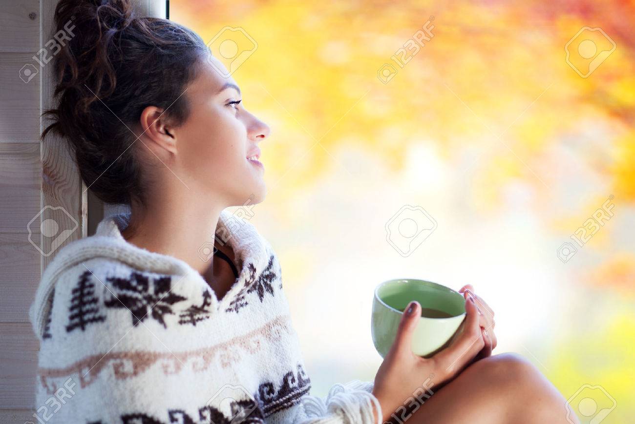Young Beaufort brunette smiling woman with cup of coffee wearing knitted nordic print poncho sitting home by the window. Blurred garden fall background. - 46287945 Young Beaufort brunette smiling woman with cup of coffee wearing knitted nordic print poncho sitting home by the window. Blurred garden fall background. - 46287945