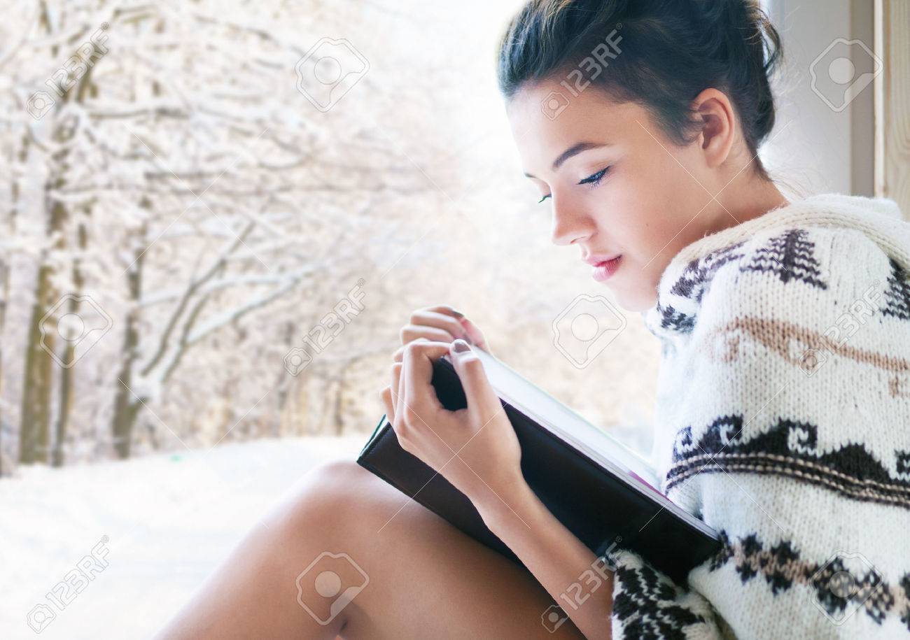 Beautiful young brunette woman reading book wearing knitted poncho sitting home by the window. Snowy winter forrest background. - 49400408 Beautiful young brunette woman reading book wearing knitted poncho sitting home by the window. Snowy winter forrest background. - 49400408