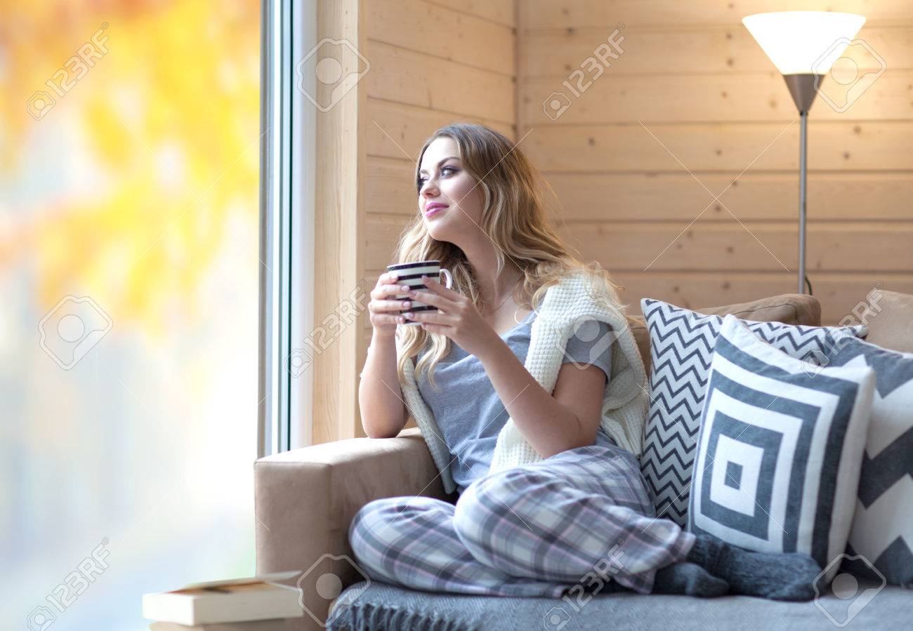 Young beautiful blonde woman with cup of coffee sitting home by the window. Autumn fall trees reflection on the glass. Lazy day off concept - 62849117 Young beautiful blonde woman with cup of coffee sitting home by the window. Autumn fall trees reflection on the glass. Lazy day off concept - 62849117