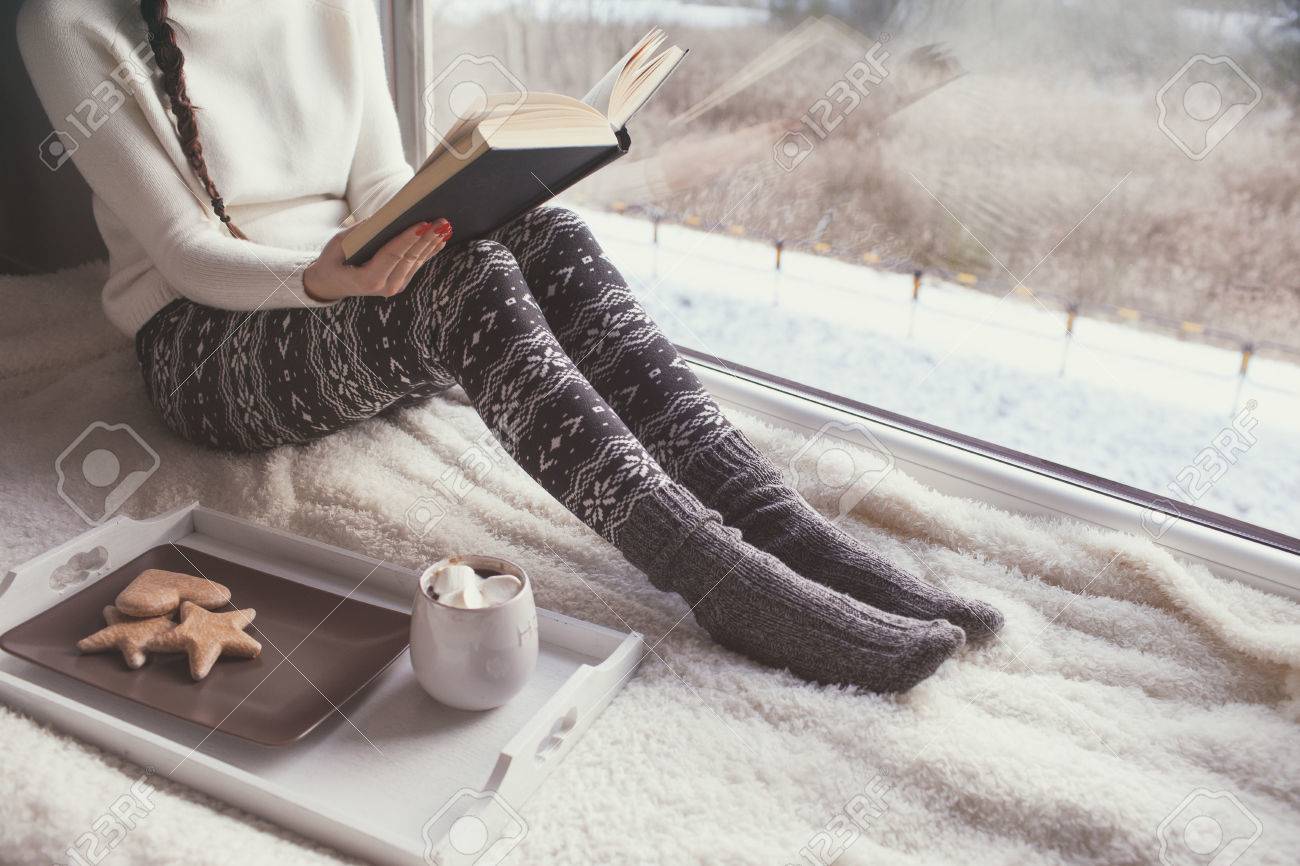 Young woman with cup of coffee and gingerbread is reading a book sitting home relaxing by the window. Snow winter home chill out concept. - 74409347 Young woman with cup of coffee and gingerbread is reading a book sitting home relaxing by the window. Snow winter home chill out concept. - 74409347