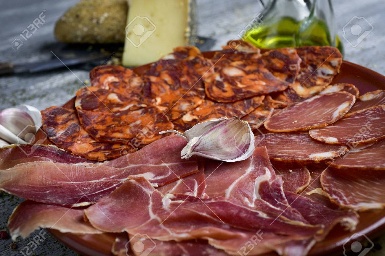 a plate with an assortment of different spanish cold meats as chorizo, cured pork tenderloin and serrano ham, and a piece of manchego cheese, bread and a cruet with olive oil in the background - 73006043 a plate with an assortment of different spanish cold meats as chorizo, cured pork tenderloin and serrano ham, and a piece of manchego cheese, bread and a cruet with olive oil in the background - 73006043