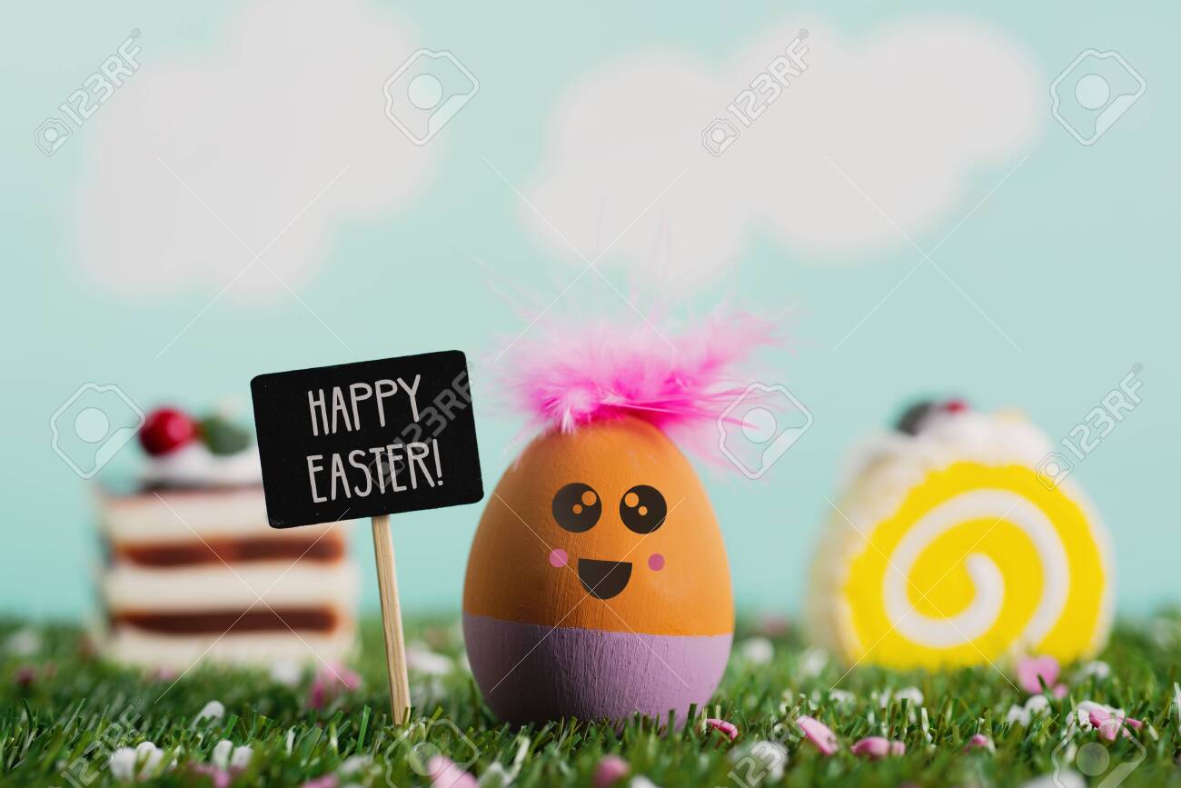 a handmade easter egg, with a cute face, on the grass, next to a black signboard with the text happy easter written in it, some pieces of colorful cakes and a sky with clouds in the background - 120706258 a handmade easter egg, with a cute face, on the grass, next to a black signboard with the text happy easter written in it, some pieces of colorful cakes and a sky with clouds in the background - 120706258