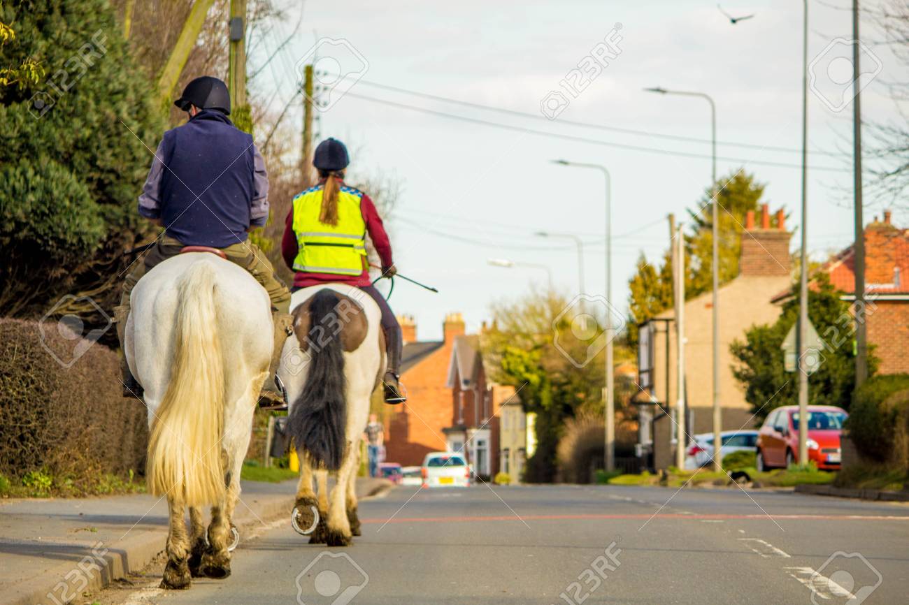 Two horse riders travel through a small village, on the main road. - 99084210 Two horse riders travel through a small village, on the main road. - 99084210