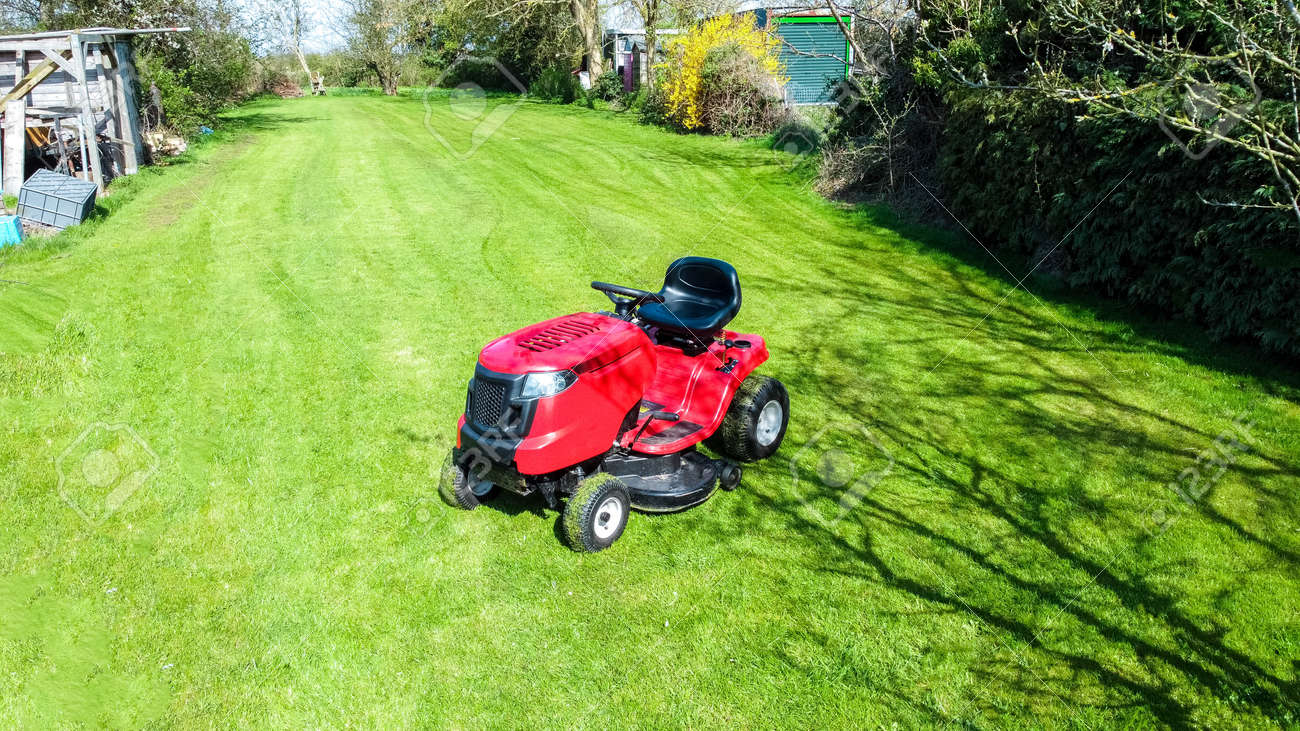 A view of a large ride along lawn mower cutting the grass of a large garden, at the back of a small property. The sun shining above during spring time. - 168149661 A view of a large ride along lawn mower cutting the grass of a large garden, at the back of a small property. The sun shining above during spring time. - 168149661