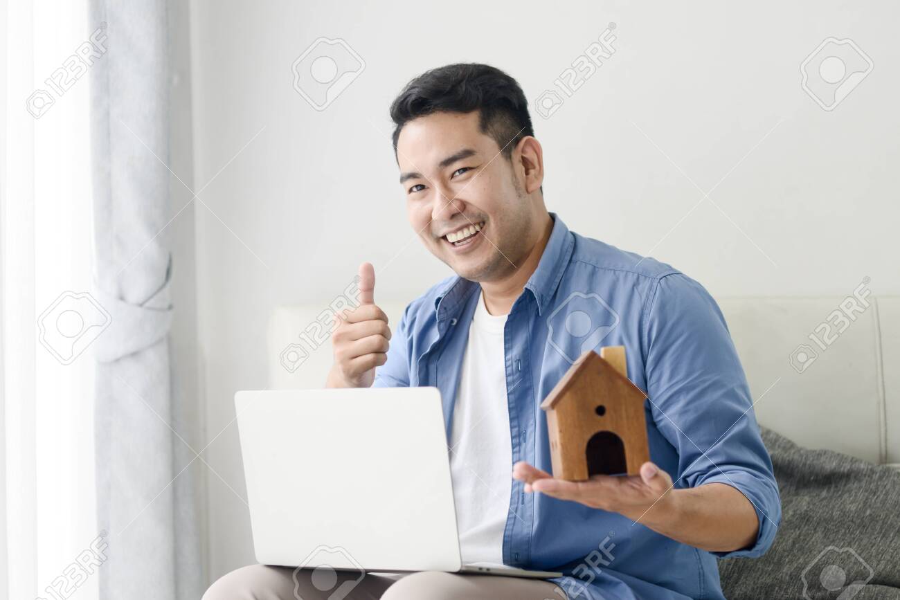 Asian man holding house in his hand and using laptop, real estate concept. - 129901049 Asian man holding house in his hand and using laptop, real estate concept. - 129901049