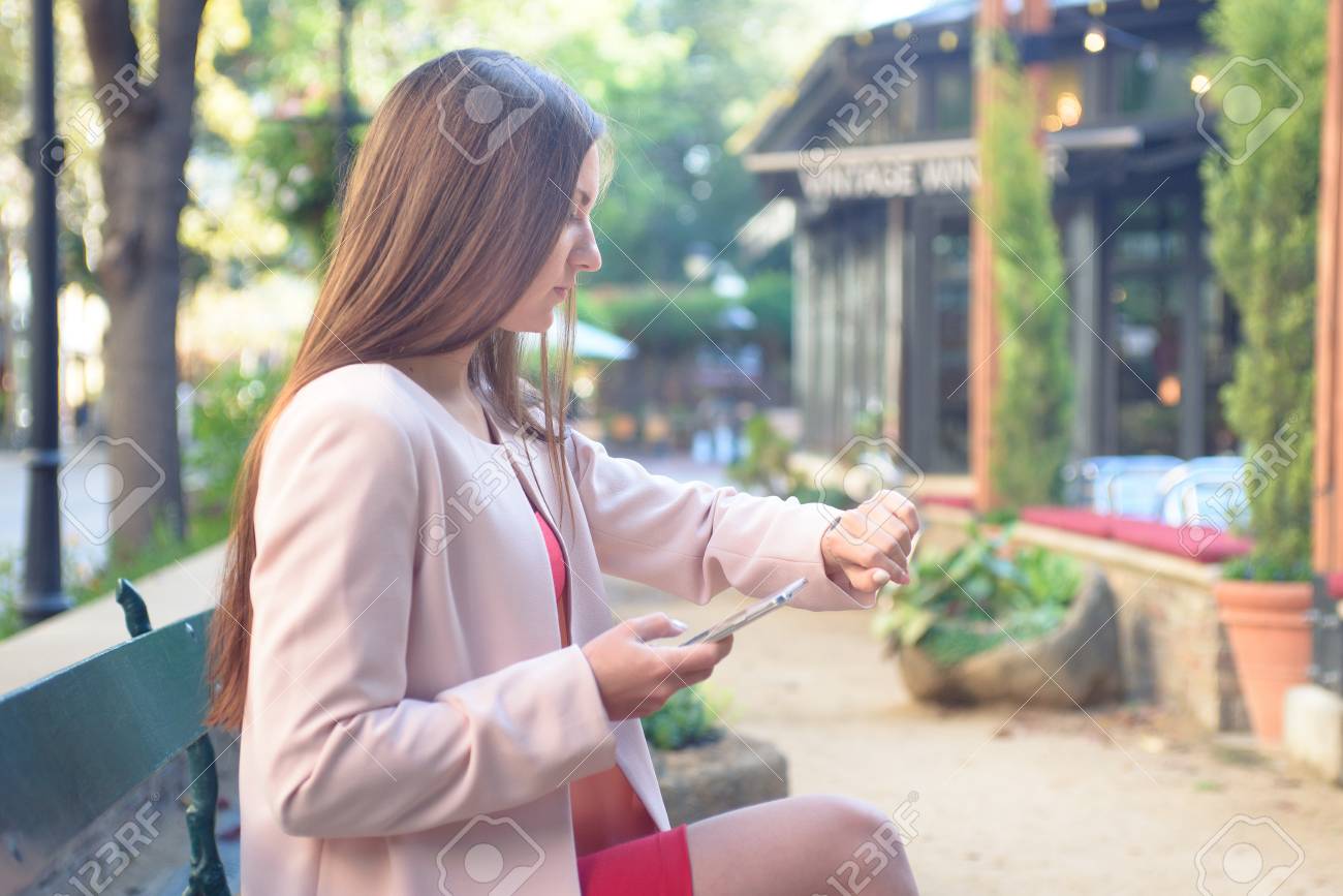 Young attractive woman, sitting alone on a bench outside, impatiently checking time on her wrist watch, holding cell phone. - 71050104 Young attractive woman, sitting alone on a bench outside, impatiently checking time on her wrist watch, holding cell phone. - 71050104