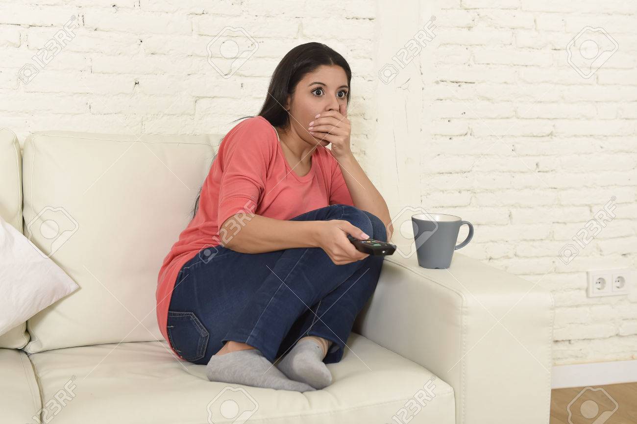 若い美しいラテン女性自宅テレビ怖いホラー映画を見たり 恐ろしいニュースの居間でソファー ソファで座って怖がってし リモート コントロールを保持している彼女の口を覆っている興奮して の写真素材 画像素材 Image