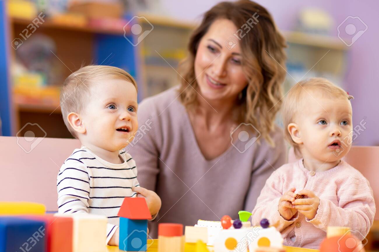 Kindergarten teacher looking after children in daycare. Nursery babies play together with developmental toys. - 138983907 Kindergarten teacher looking after children in daycare. Nursery babies play together with developmental toys. - 138983907