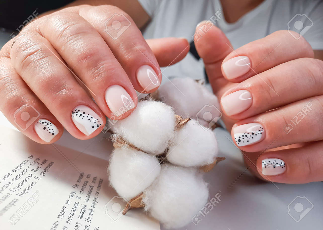 Soft pink gel polish with a design of black dots on the background of the text of the book. Women's hands with pastel colored nails hold a sprig of cotton. Milk manicure with black and white design. - 159836169 Soft pink gel polish with a design of black dots on the background of the text of the book. Women's hands with pastel colored nails hold a sprig of cotton. Milk manicure with black and white design. - 159836169