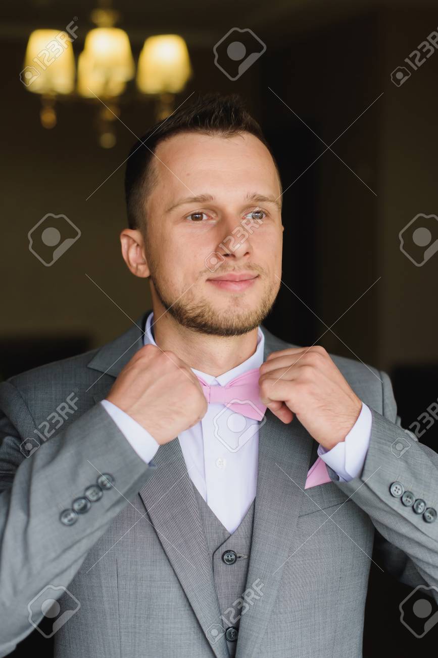 Hombre Joven Elegante Que Se Viste Para La Celebracion De La Boda Apuesto Novio Vestido Con Traje Gris Moderno Camisa Blanca Y Pajarita Rosa Preparandose Para El Evento El Hombre Ajusta Su
