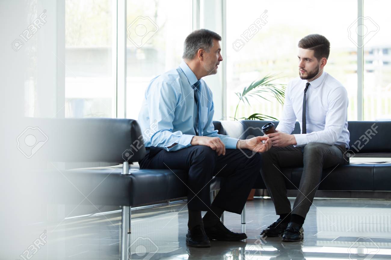 Mature businessman using a digital tablet to discuss information with a younger colleague in a modern business lounge. - 117097432 Mature businessman using a digital tablet to discuss information with a younger colleague in a modern business lounge. - 117097432