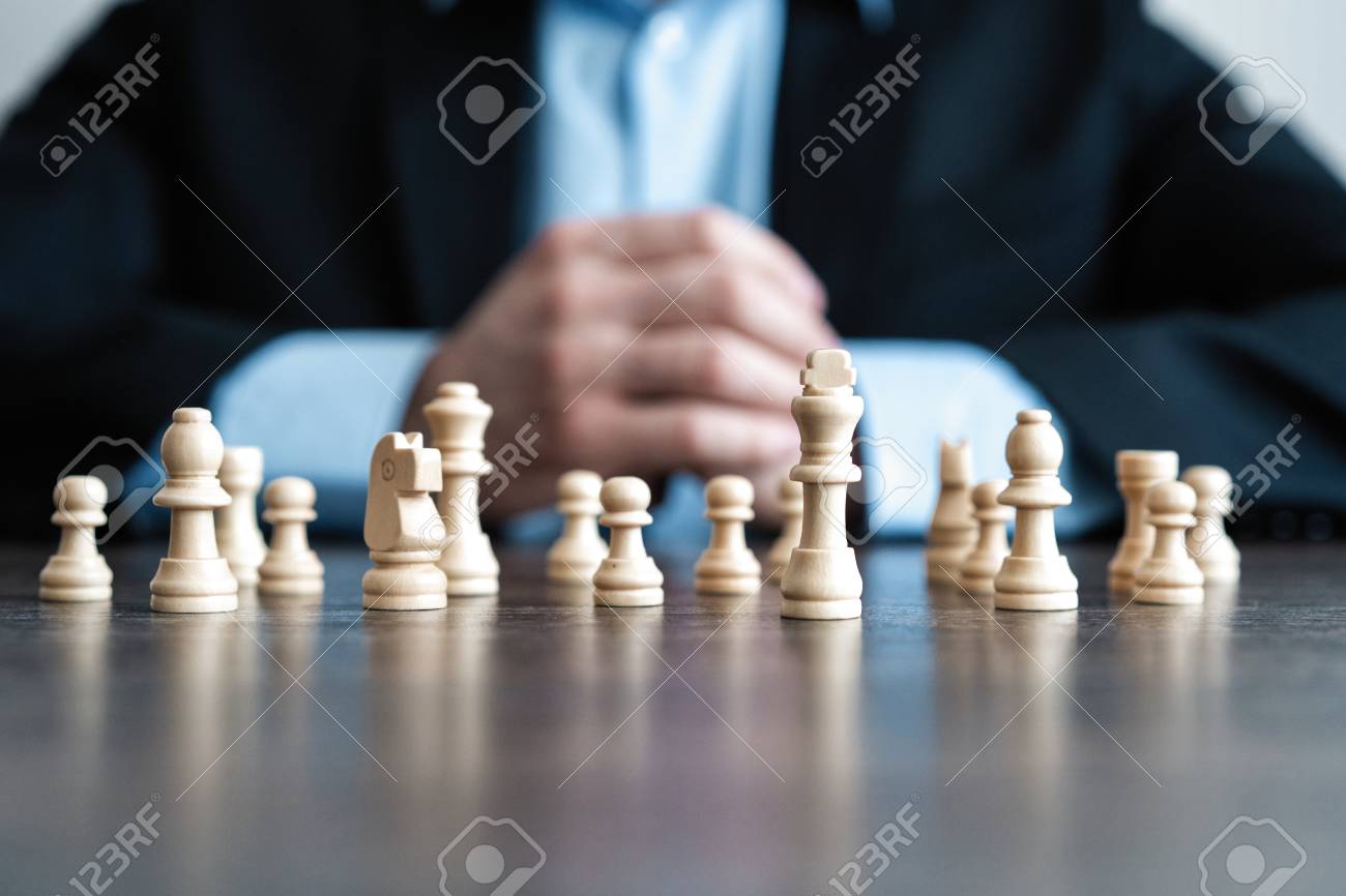 Businessman with clasped hands planning strategy with chess figures on table. Strategy, leadership and teamwork concept. - 119770100 Businessman with clasped hands planning strategy with chess figures on table. Strategy, leadership and teamwork concept. - 119770100