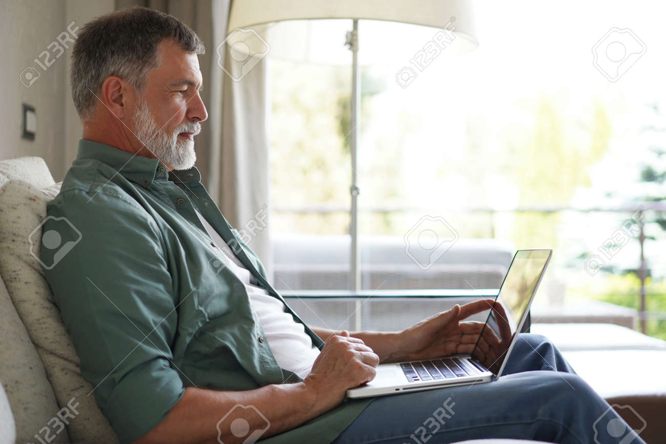 Portrait of happy mature man in casual clothes using laptop lying on sofa in house. - 171563191 Portrait of happy mature man in casual clothes using laptop lying on sofa in house. - 171563191