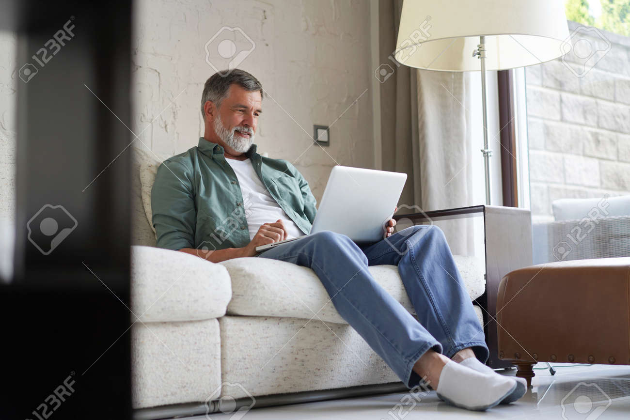 Portrait of happy mature man in casual clothes using laptop lying on sofa in house. - 171563456 Portrait of happy mature man in casual clothes using laptop lying on sofa in house. - 171563456