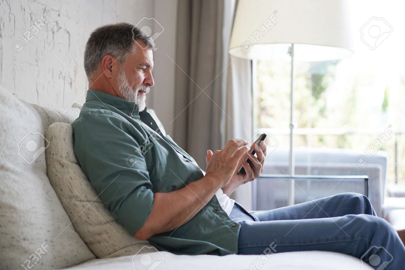 Smiling Senior Man Using Cellphone Browsing Internet Sitting On Sofa Indoor. - 171566653 Smiling Senior Man Using Cellphone Browsing Internet Sitting On Sofa Indoor. - 171566653