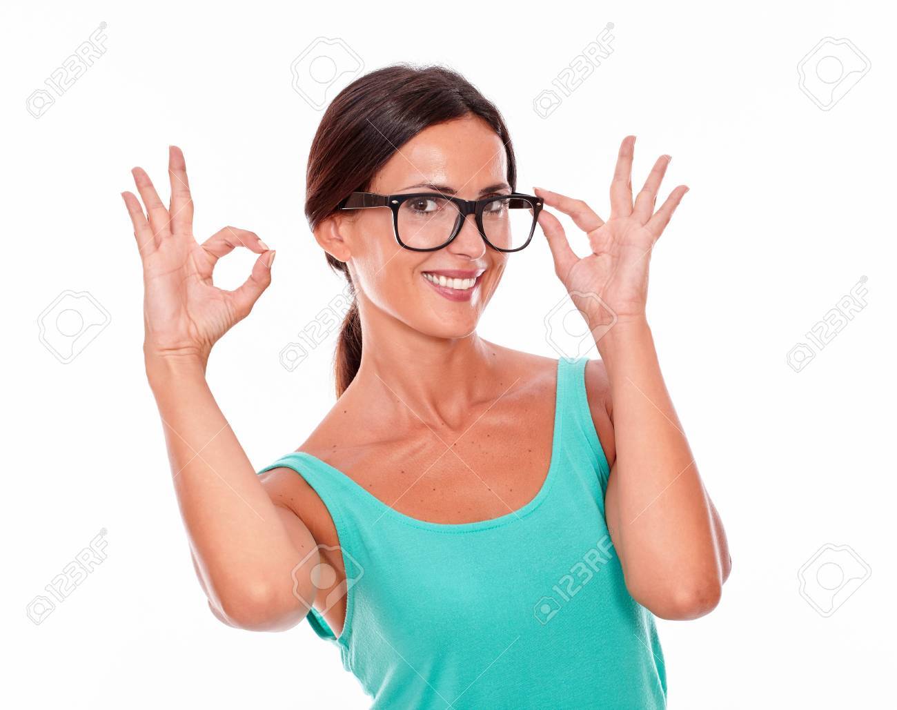 カメラでニコニコと片方の手で Ok サインを示し 他の着ている白い背景に緑のタンクトップで結ばれる彼女の長い髪を持つ彼女のメガネに触れて満足している大人の女性 の写真素材 画像素材 Image