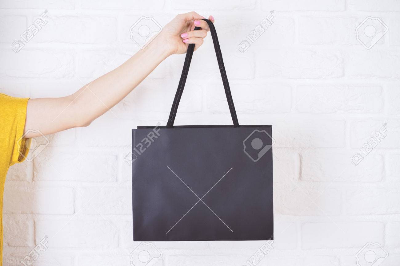 Closeup of female hand holding black shopping bag on white brick background. - 54889720 Closeup of female hand holding black shopping bag on white brick background. - 54889720