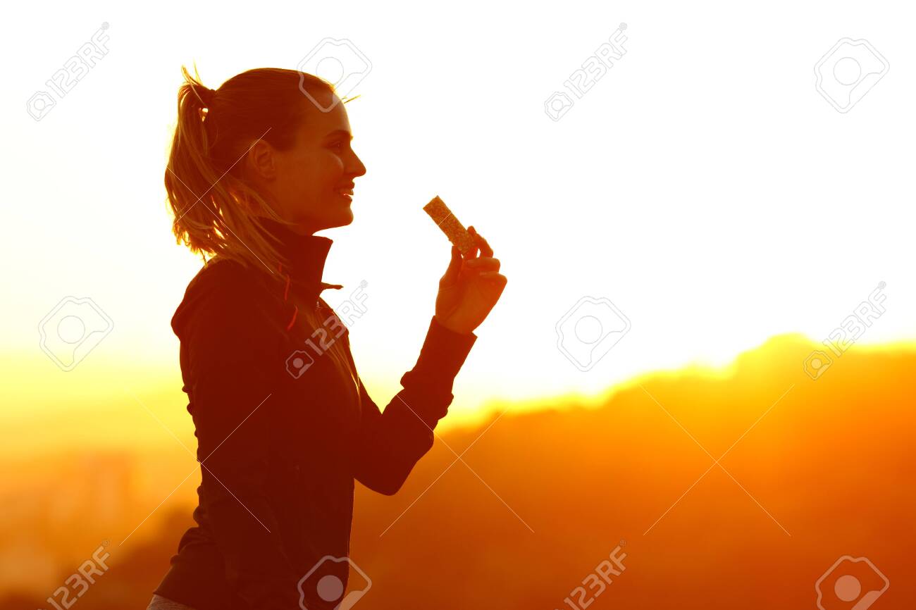 Silhouette of runner woman eating energy bar after running at sunset in the mountain - 140301623 Silhouette of runner woman eating energy bar after running at sunset in the mountain - 140301623