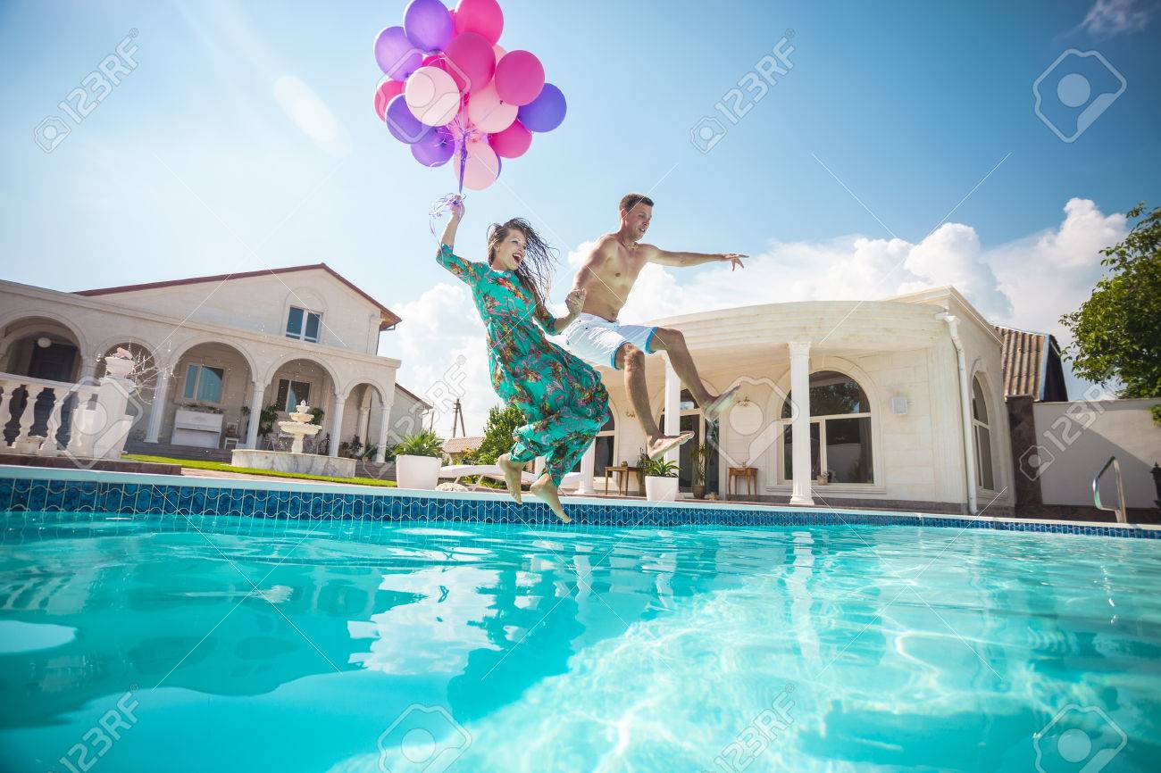 Happy young couple jumping into the pool while holding a bunch of balloons - 48996569 Happy young couple jumping into the pool while holding a bunch of balloons - 48996569