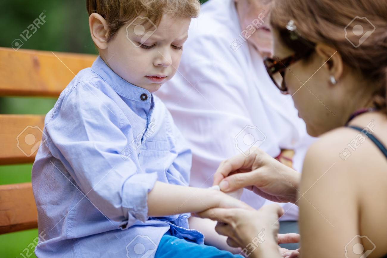 Mother applying band aid on scratch on son's arm while sitting on bench in park. Senior grandmother sitting beside on bench. - 81557890 Mother applying band aid on scratch on son's arm while sitting on bench in park. Senior grandmother sitting beside on bench. - 81557890