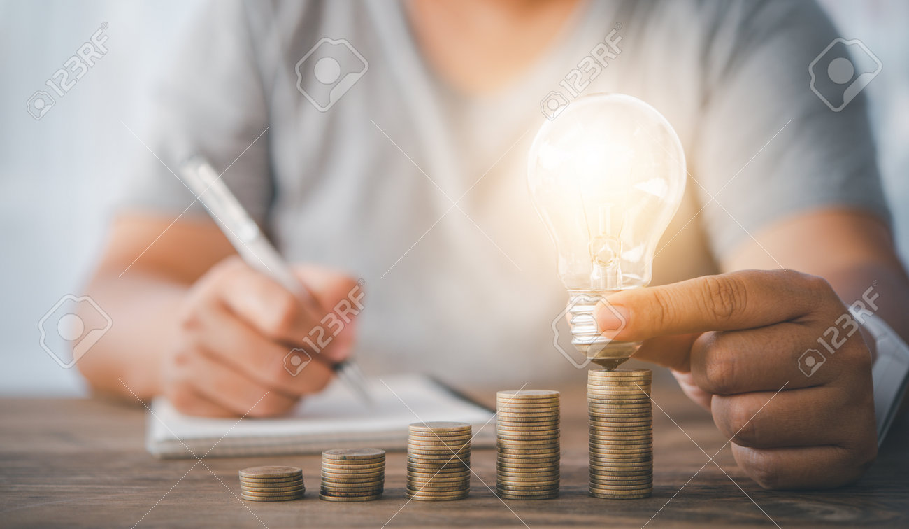 woman's hand holding a light bulb on a coin lying on a desk.Energy saving ideas to cut home expenses. - 163301659 woman's hand holding a light bulb on a coin lying on a desk.Energy saving ideas to cut home expenses. - 163301659