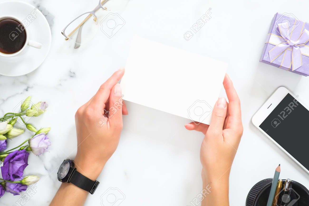 Female hands holding blank paper card mockup over modern home office desk workspace with stationery, cup of coffee, flowers bouquet, glasses, smartphone. Flat lay, top view - 159015030 Female hands holding blank paper card mockup over modern home office desk workspace with stationery, cup of coffee, flowers bouquet, glasses, smartphone. Flat lay, top view - 159015030