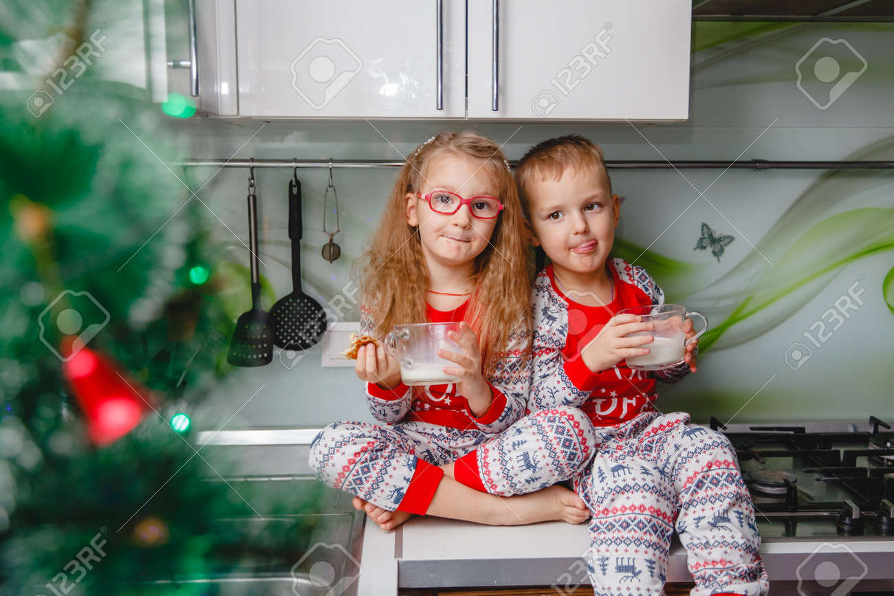 Little brother and sister sit on a Christmas-decorated kitchen table in pajamas and drink milk. Girl with glasses - 159148180 Little brother and sister sit on a Christmas-decorated kitchen table in pajamas and drink milk. Girl with glasses - 159148180