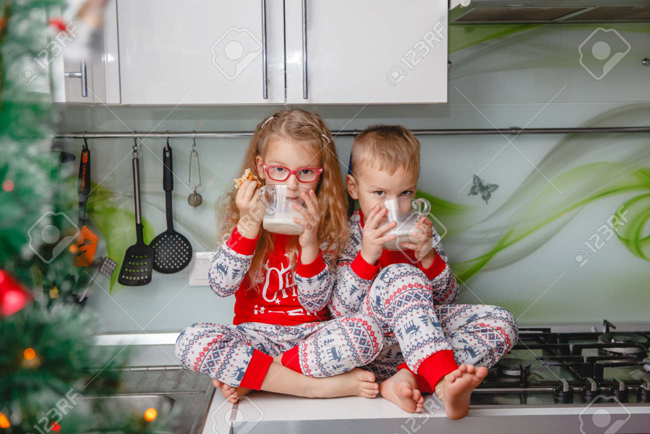 Little brother and sister sit on a Christmas-decorated kitchen table in pajamas and drink milk. Girl with glasses - 159146734 Little brother and sister sit on a Christmas-decorated kitchen table in pajamas and drink milk. Girl with glasses - 159146734