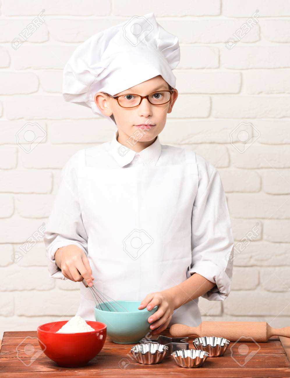 白い制服と帽子にメガネ麺棒とカラフルなボウルを持つテーブルの近くに立って レンガ壁の背景に料理を真剣な顔で少年の小さなかわいい料理人シェフ の写真素材 画像素材 Image