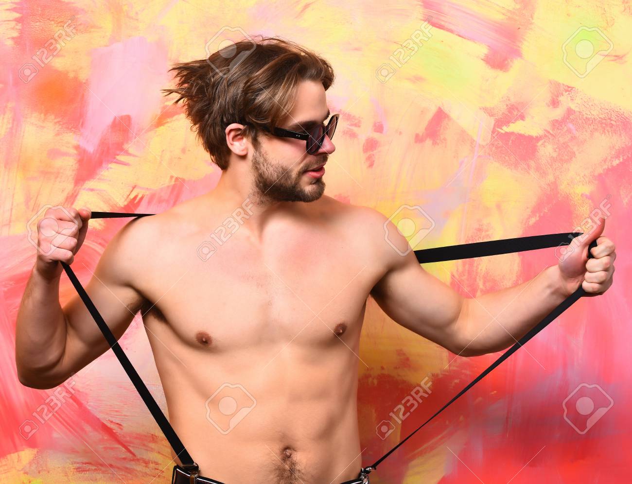 口ひげを持つ白人マッチョ男パンツ サスペンダーとカラフルなスタジオの背景にサングラスで筋肉の胴体に六つのパックと Abs を示しています の写真素材 画像素材 Image