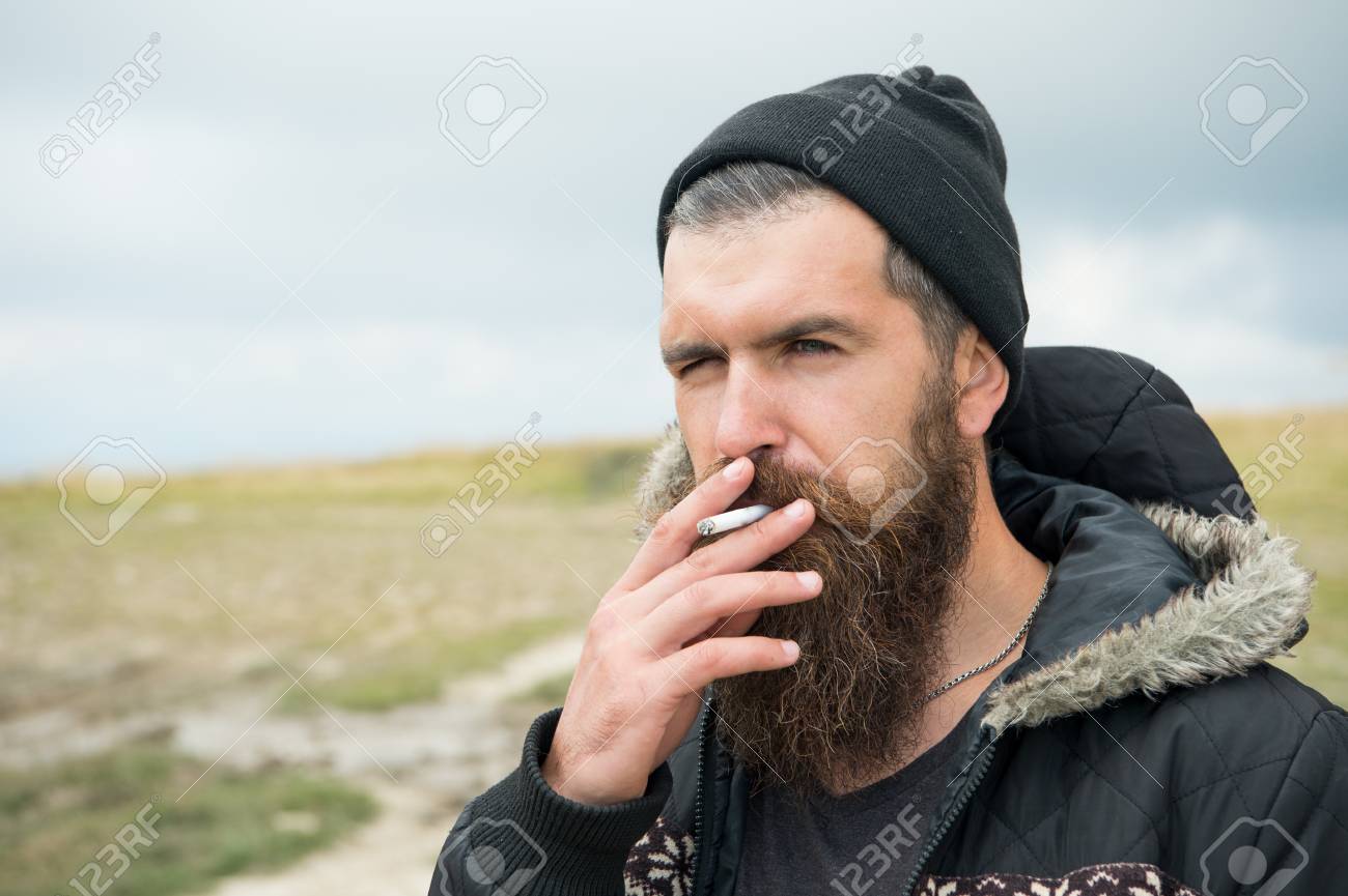 Homme qui fume. jeune homme barbu hipster fumer cigarette en chapeau et veste à la montagne et ciel nuageux sur fond naturel, espace copie