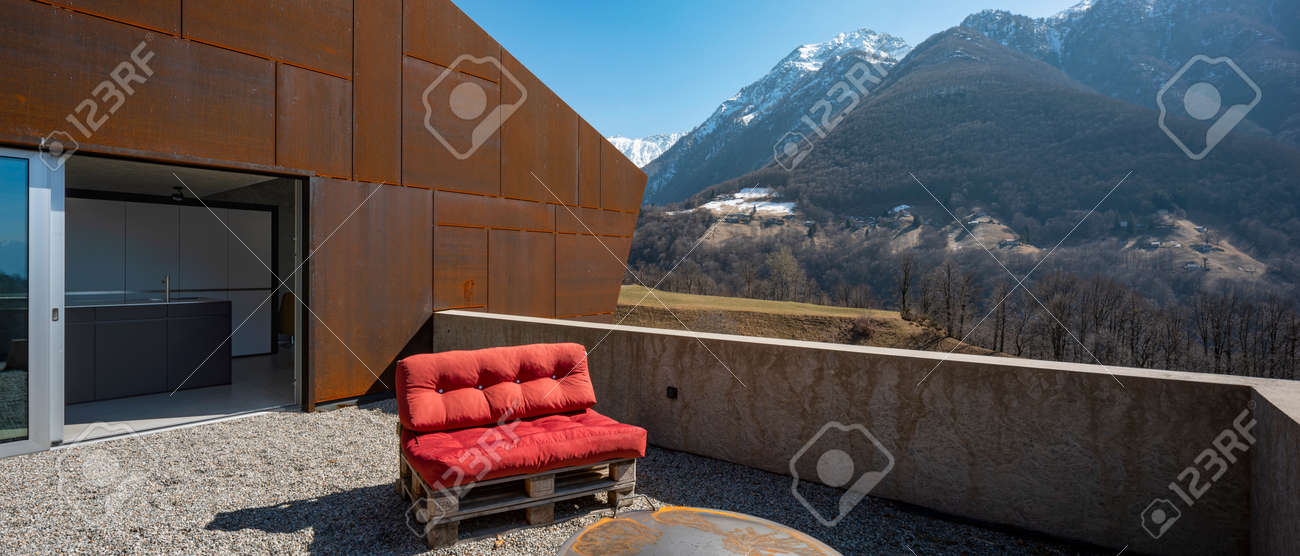 Modern house exteriors with veranda. Red sofa overlooking the mountains of Switzerland. Concept, nobody inside - 166390035 Modern house exteriors with veranda. Red sofa overlooking the mountains of Switzerland. Concept, nobody inside - 166390035