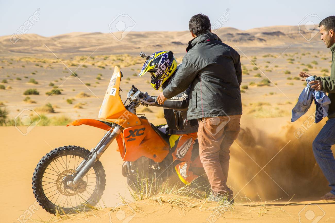 Merzouga, Morocco - February 25, 2016: A motorbike rider churns up sand as he tries to get his orange bike going in the Sahara Desert. Two men are coming to his assistance. In the distance you can see rougher terrain; sand with tufts of grass and hills - 93588231 Merzouga, Morocco - February 25, 2016: A motorbike rider churns up sand as he tries to get his orange bike going in the Sahara Desert. Two men are coming to his assistance. In the distance you can see rougher terrain; sand with tufts of grass and hills - 93588231