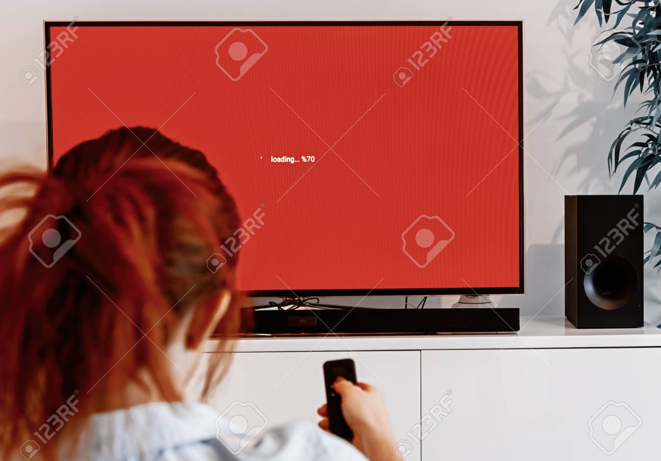 Redhead woman sitting in her living room and holding a TV remote control in front of a screen waiting message while loading an application on her connected TV - 115162264 Redhead woman sitting in her living room and holding a TV remote control in front of a screen waiting message while loading an application on her connected TV - 115162264