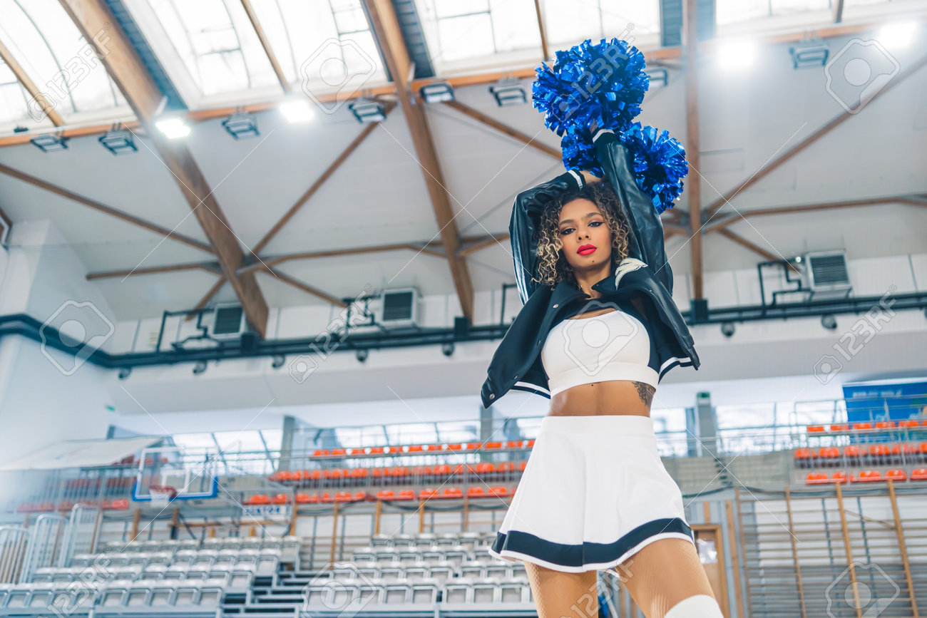 チア jk 頭上に青いポンポンを持つ革のジャケットを着たチアリーダーの底面図。背景にぼかしたバスケットボールコート。高品質の写真の写真素材・画像素材 Image  199709741