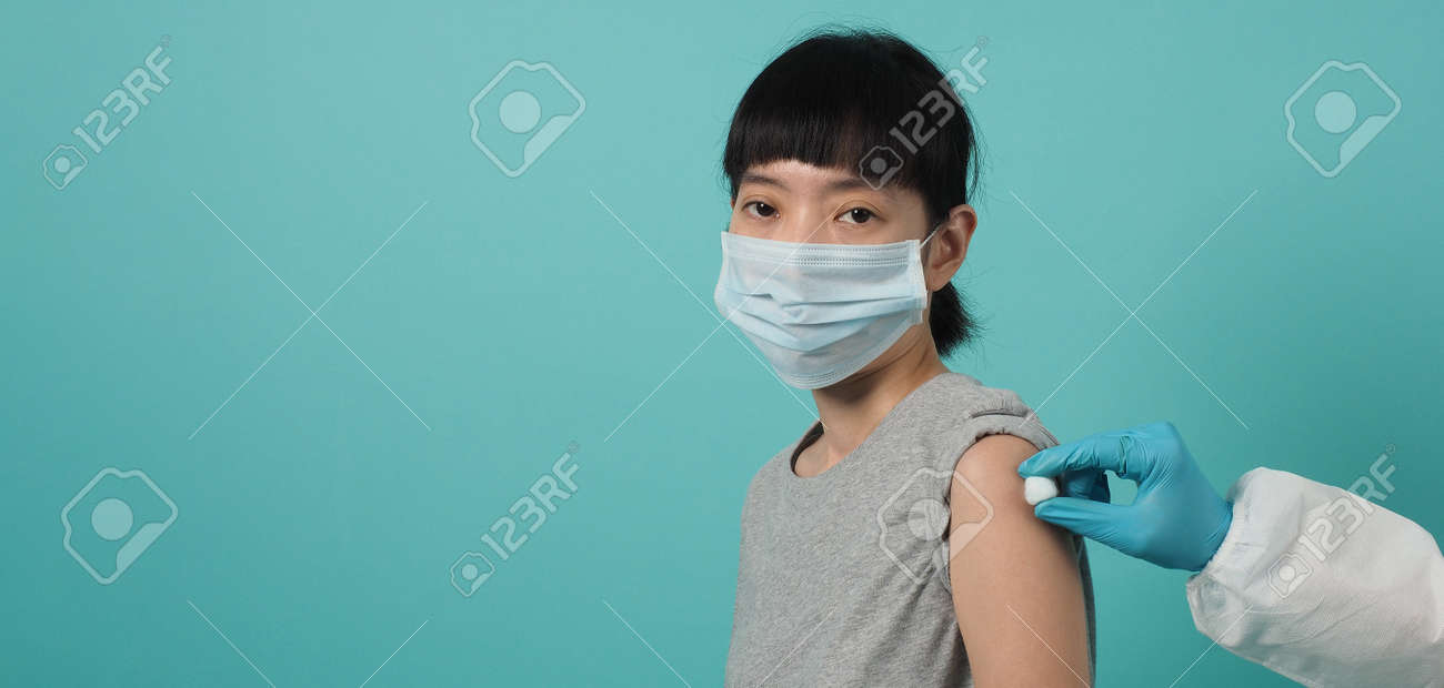 Vaccine Concept. Woman waiting for coronavirus vaccine injection by doctor. Asian woman with medical mask open shoulder and upper arm on blue green background. Waiting for vaccination. - 168300451 Vaccine Concept. Woman waiting for coronavirus vaccine injection by doctor. Asian woman with medical mask open shoulder and upper arm on blue green background. Waiting for vaccination. - 168300451